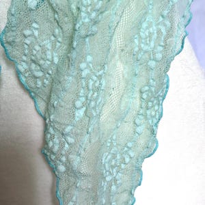 May include: A delicate, light blue scarf with a textured, floral-like pattern. The edges are trimmed with a contrasting turquoise border. The scarf is draped over a white form, showcasing its airy, lightweight design.