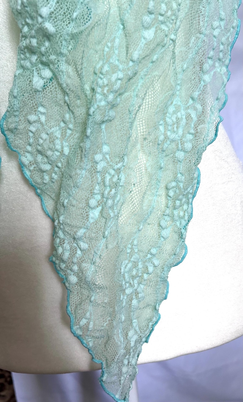 May include: A delicate, light blue scarf with a textured, floral-like pattern. The edges are trimmed with a contrasting turquoise border. The scarf is draped over a white form, showcasing its airy, lightweight design.