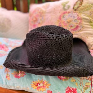 May include: A black woven sun hat with a wide brim. The hat is resting on a floral patterned cushion with a light blue background. The hat's crown is cylindrical.