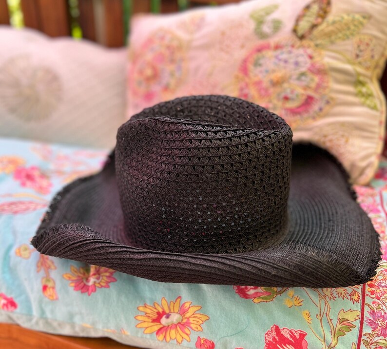 May include: A black woven sun hat with a wide brim. The hat is resting on a floral patterned cushion with a light blue background. The hat's crown is cylindrical.