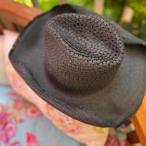May include: A dark grey woven cowboy hat with a wide brim. The hat has a perforated crown and is made of a textured material. The hat is resting on a floral patterned surface.