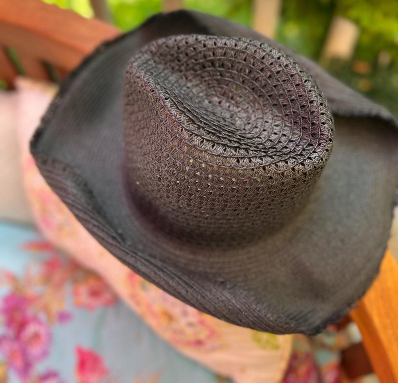 May include: A dark grey woven cowboy hat with a wide brim. The hat has a perforated crown and is made of a textured material. The hat is resting on a floral patterned surface.