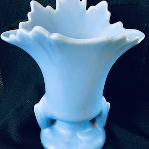 May include: A light blue ceramic vase with a ruffled rim and a flared body. The vase has two small handles near the base and a rounded bottom. The vase is approximately 6 inches tall.