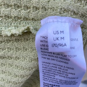 Puede incluir: Una etiqueta blanca con texto negro que detalla el tama&ntilde;o y las instrucciones de cuidado de una prenda. La etiqueta dice "US M, UK M, 170/96A, Suave, Lavar a m&aacute;quina con agua fr&iacute;a, ciclo delicado, con colores similares, usar solo blanqueador sin cloro si es necesario, remodelar y secar plano, planchar a baja temperatura si es necesario, se puede limpiar en seco."