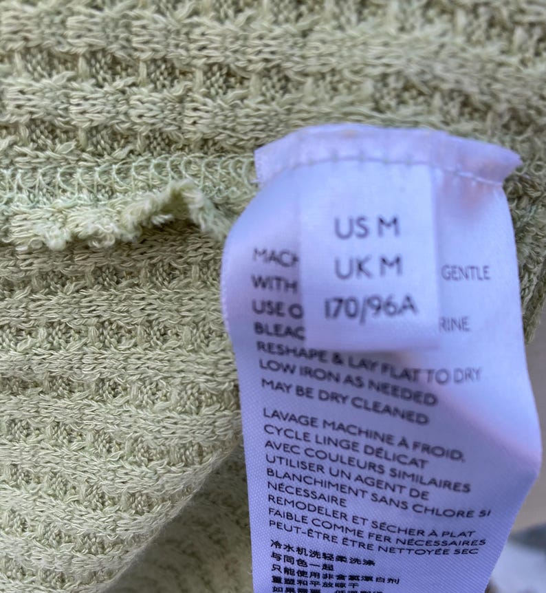 Puede incluir: Una etiqueta blanca con texto negro que detalla el tama&ntilde;o y las instrucciones de cuidado de una prenda. La etiqueta dice "US M, UK M, 170/96A, Suave, Lavar a m&aacute;quina con agua fr&iacute;a, ciclo delicado, con colores similares, usar solo blanqueador sin cloro si es necesario, remodelar y secar plano, planchar a baja temperatura si es necesario, se puede limpiar en seco."