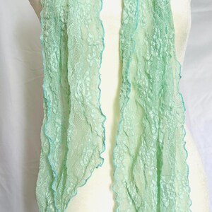 May include: A delicate, mint green lace scarf with a scalloped edge and a contrasting light blue trim. The scarf is draped over a mannequin, showcasing its flowing, lightweight design. This accessory adds a touch of elegance.
