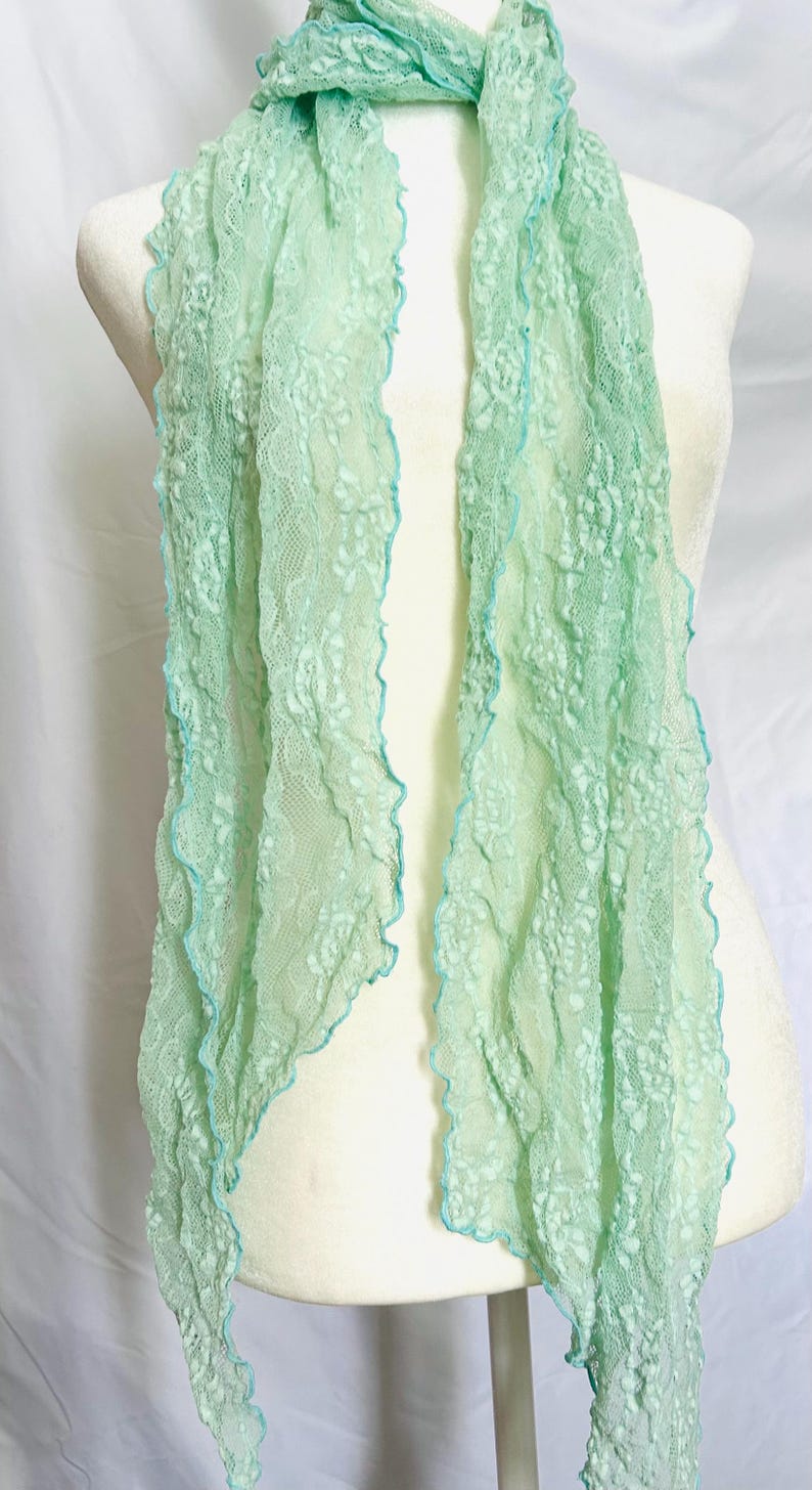 May include: A delicate, mint green lace scarf with a scalloped edge and a contrasting light blue trim. The scarf is draped over a mannequin, showcasing its flowing, lightweight design. This accessory adds a touch of elegance.
