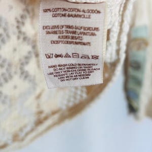 May include: Close-up of a cream-colored garment with a lace design and a printed tag. The tag includes washing instructions and fabric composition details. The garment is made of 100% cotton.