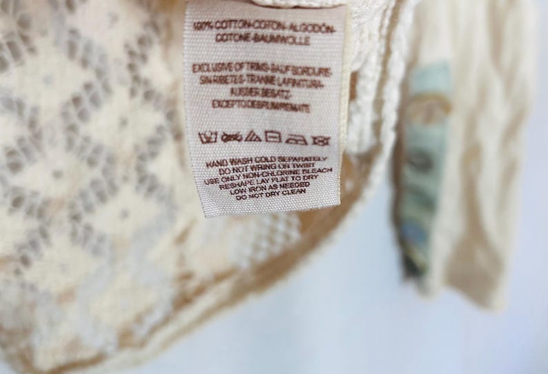 May include: Close-up of a cream-colored garment with a lace design and a printed tag. The tag includes washing instructions and fabric composition details. The garment is made of 100% cotton.