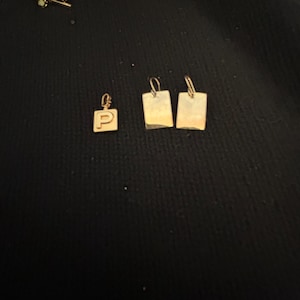 May include: Gold-toned jewelry pieces, including a pendant with the letter "P" and two rectangular earrings. The items are displayed on a dark surface, showcasing their metallic sheen and simple design.