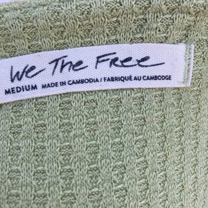 Puede incluir: Una blusa tejida verde con una etiqueta blanca que dice "We The Free" y "MEDIUM MADE IN CAMBODIA / FABRIQU&Eacute; AU CAMBODGE".