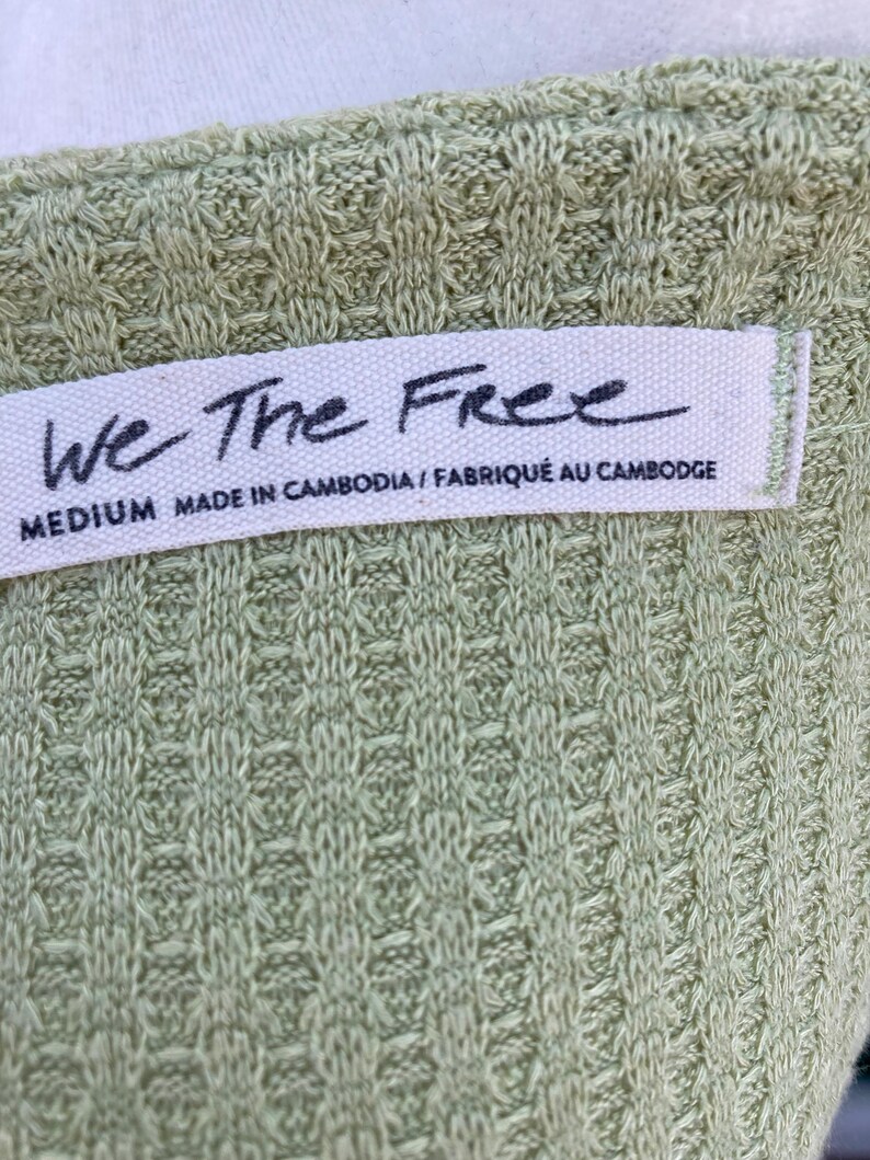 Puede incluir: Una blusa tejida verde con una etiqueta blanca que dice "We The Free" y "MEDIUM MADE IN CAMBODIA / FABRIQU&Eacute; AU CAMBODGE".