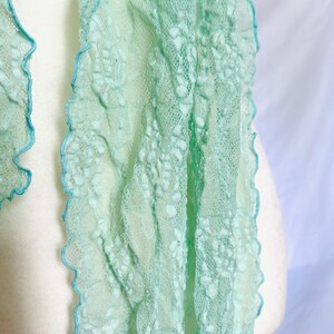 May include: A delicate, mint green scarf with a textured, open weave design. The edges are trimmed with a scalloped, light blue border. The scarf is draped over a white form, showcasing its airy and lightweight quality.