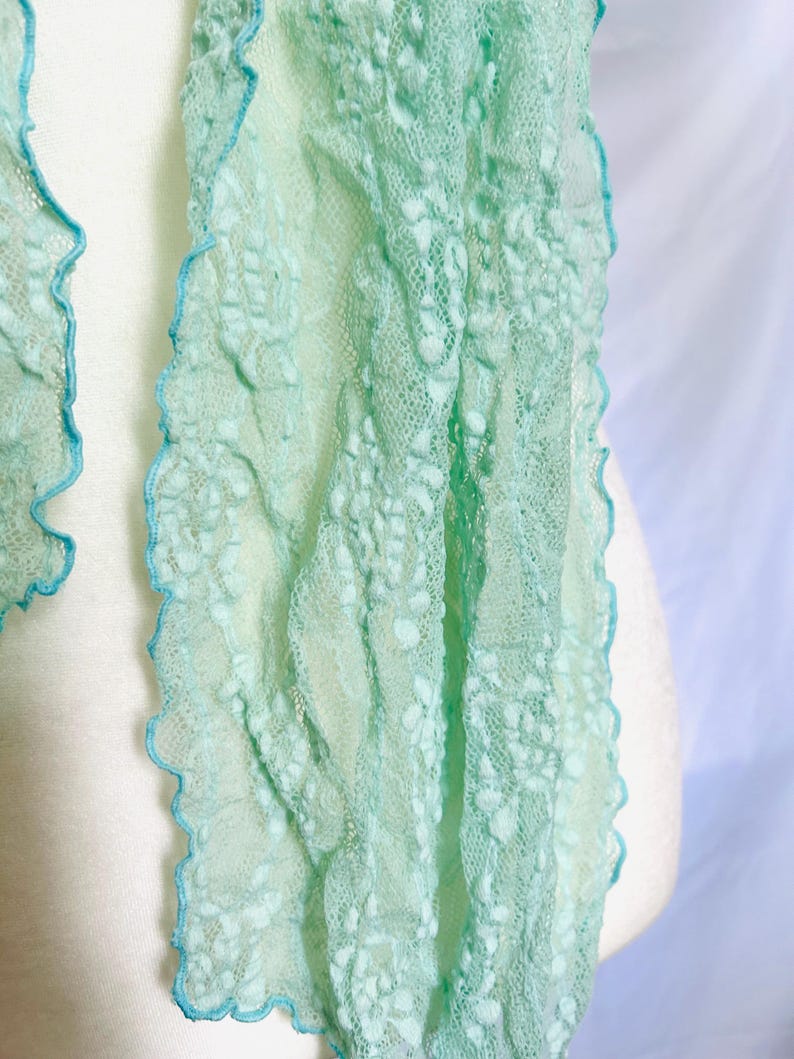 May include: A delicate, mint green scarf with a textured, open weave design. The edges are trimmed with a scalloped, light blue border. The scarf is draped over a white form, showcasing its airy and lightweight quality.