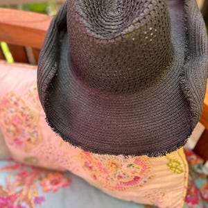May include: A black sun hat with a wide brim and a textured weave. The hat has a frayed edge and is resting on a floral patterned pillow. The hat's crown is circular and the brim is wide, providing ample shade.