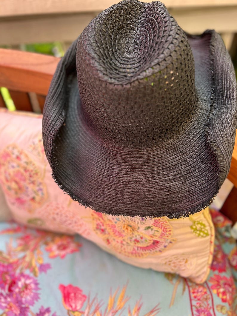 May include: A black sun hat with a wide brim and a textured weave. The hat has a frayed edge and is resting on a floral patterned pillow. The hat's crown is circular and the brim is wide, providing ample shade.