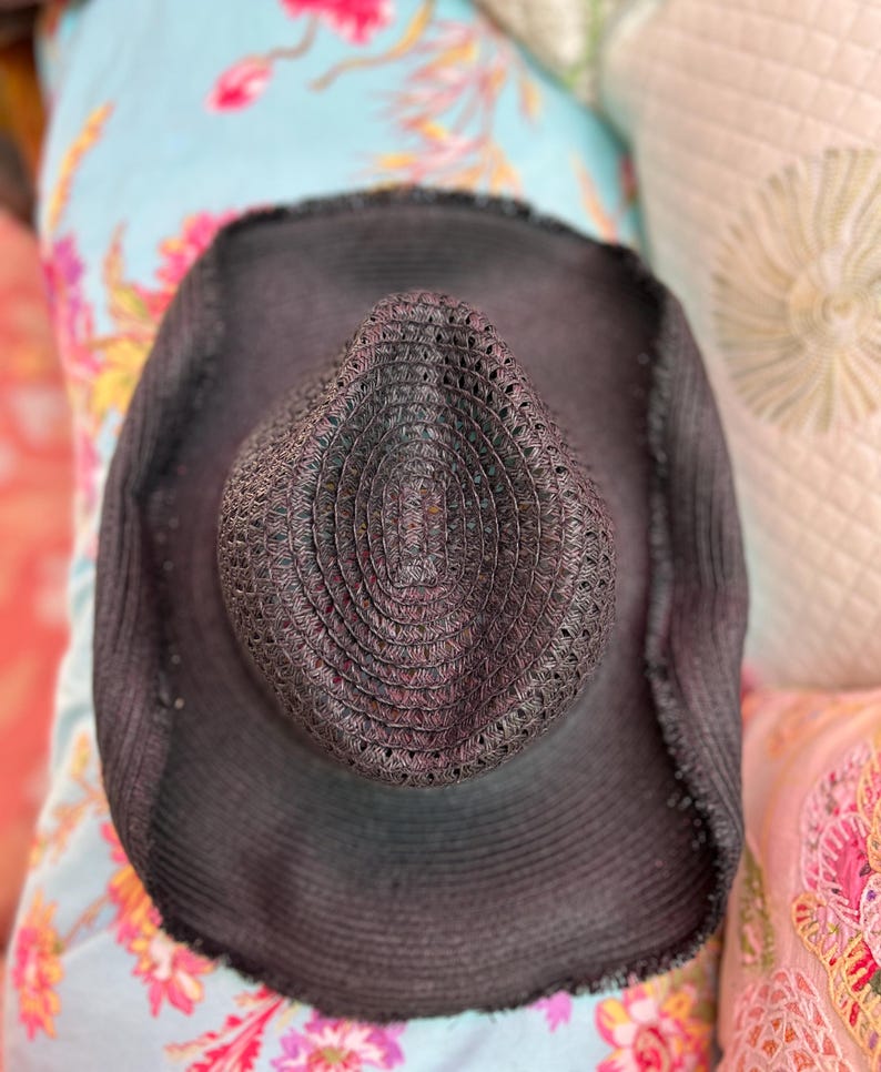 May include: A dark grey woven sun hat with a wide brim. The hat has a textured crown and a slightly frayed edge. The hat is resting on a floral patterned surface.