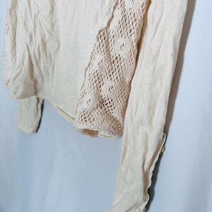 May include: A light beige long-sleeved top with a crochet panel on the right side. The top is made of a soft, lightweight fabric. The crochet panel adds a decorative touch to the garment.