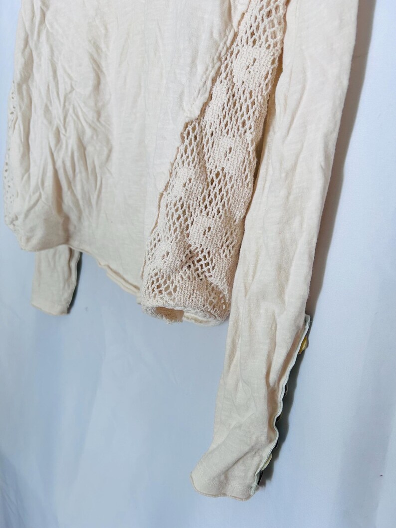 May include: A light beige long-sleeved top with a crochet panel on the right side. The top is made of a soft, lightweight fabric. The crochet panel adds a decorative touch to the garment.
