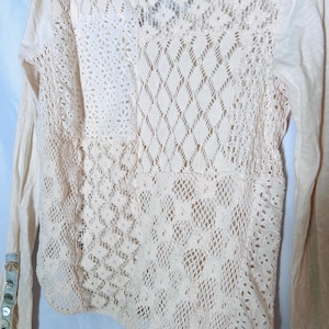 May include: Cream-colored long-sleeved shirt with intricate lace detailing on the back and sleeves. The lace features various patterns, including floral and geometric designs. The sleeves have buttoned cuffs. A casual top.