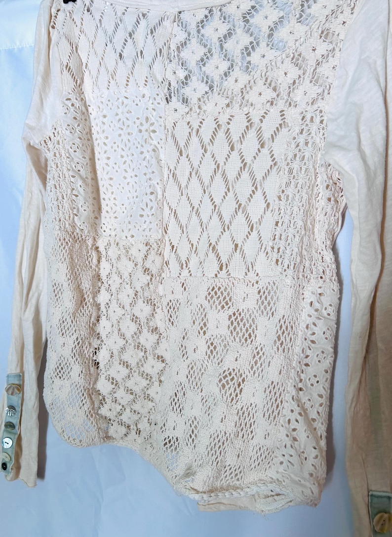 May include: Cream-colored long-sleeved shirt with intricate lace detailing on the back and sleeves. The lace features various patterns, including floral and geometric designs. The sleeves have buttoned cuffs. A casual top.