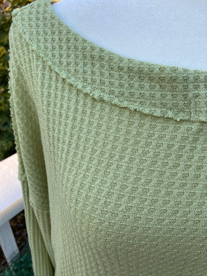 Puede incluir: Una camisa de manga larga de punto de waffle verde claro con un escote amplio y redondeado.