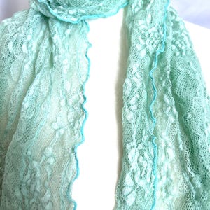 May include: A delicate, light green lace scarf with a scalloped edge trimmed in a brighter turquoise. The scarf is draped over a white form, showcasing its intricate floral pattern and airy texture. The scarf is a soft, pastel accessory.