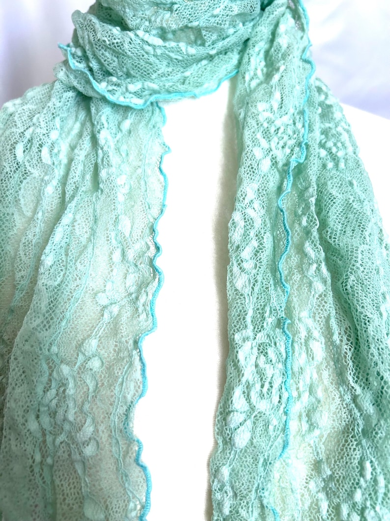 May include: A delicate, light green lace scarf with a scalloped edge trimmed in a brighter turquoise. The scarf is draped over a white form, showcasing its intricate floral pattern and airy texture. The scarf is a soft, pastel accessory.