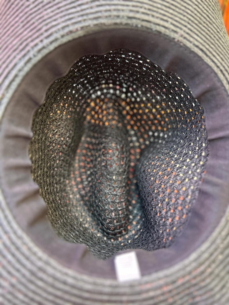 May include: A close-up of a sun hat, featuring a dark grey woven crown with small white and orange dots. The hat has a wide brim with a striped pattern in shades of grey and a dark grey interior.