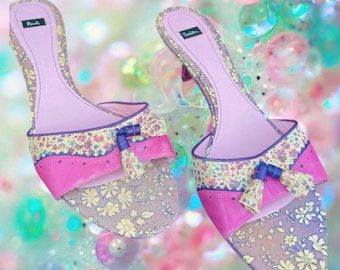 May include: A pair of open-toe mules featuring a light purple insole, a floral pattern in shades of yellow and green, and a vibrant pink leather band across the front. Each shoe is accented with a small decorative tassel and a purple trim.