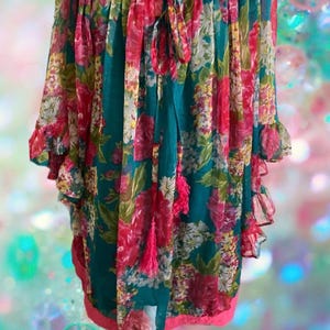 May include: A teal blouse featuring a vibrant floral pattern in shades of pink, white, and yellow. The garment has a gathered off-the-shoulder neckline with a drawstring tie, ruffled three-quarter length sleeves, and a solid pink hemline.
