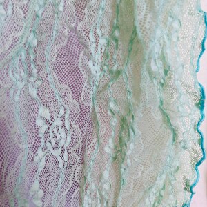 May include: Close-up of a delicate, light green lace fabric with intricate floral patterns. The lace has a scalloped edge trimmed with a darker teal color. The background is a soft purple, highlighting the detailed texture of the lace.