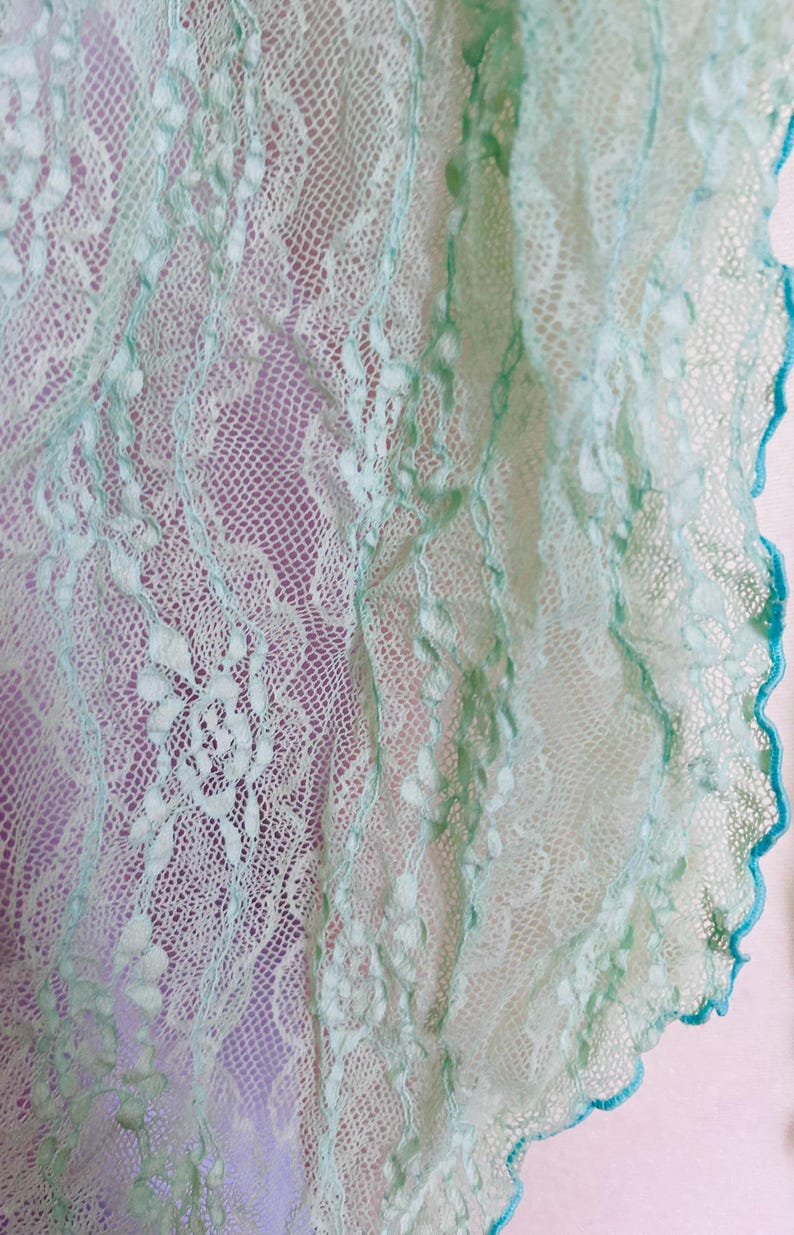 May include: Close-up of a delicate, light green lace fabric with intricate floral patterns. The lace has a scalloped edge trimmed with a darker teal color. The background is a soft purple, highlighting the detailed texture of the lace.