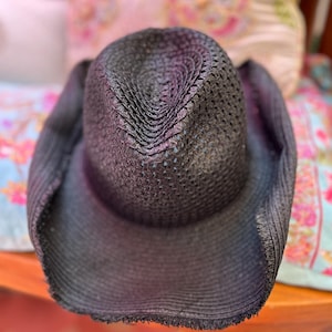 May include: A black woven cowboy hat with a wide brim. The hat has a textured, open weave design and a slightly distressed edge. The hat is resting on a wooden surface with colourful cushions in the background.
