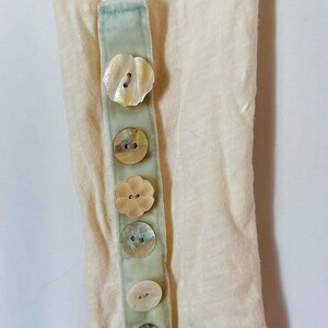 May include: Close-up of a cream-colored sleeve with a light green fabric strip. The strip is adorned with five iridescent shell buttons in various shapes. The sleeve's fabric has a slightly textured appearance.