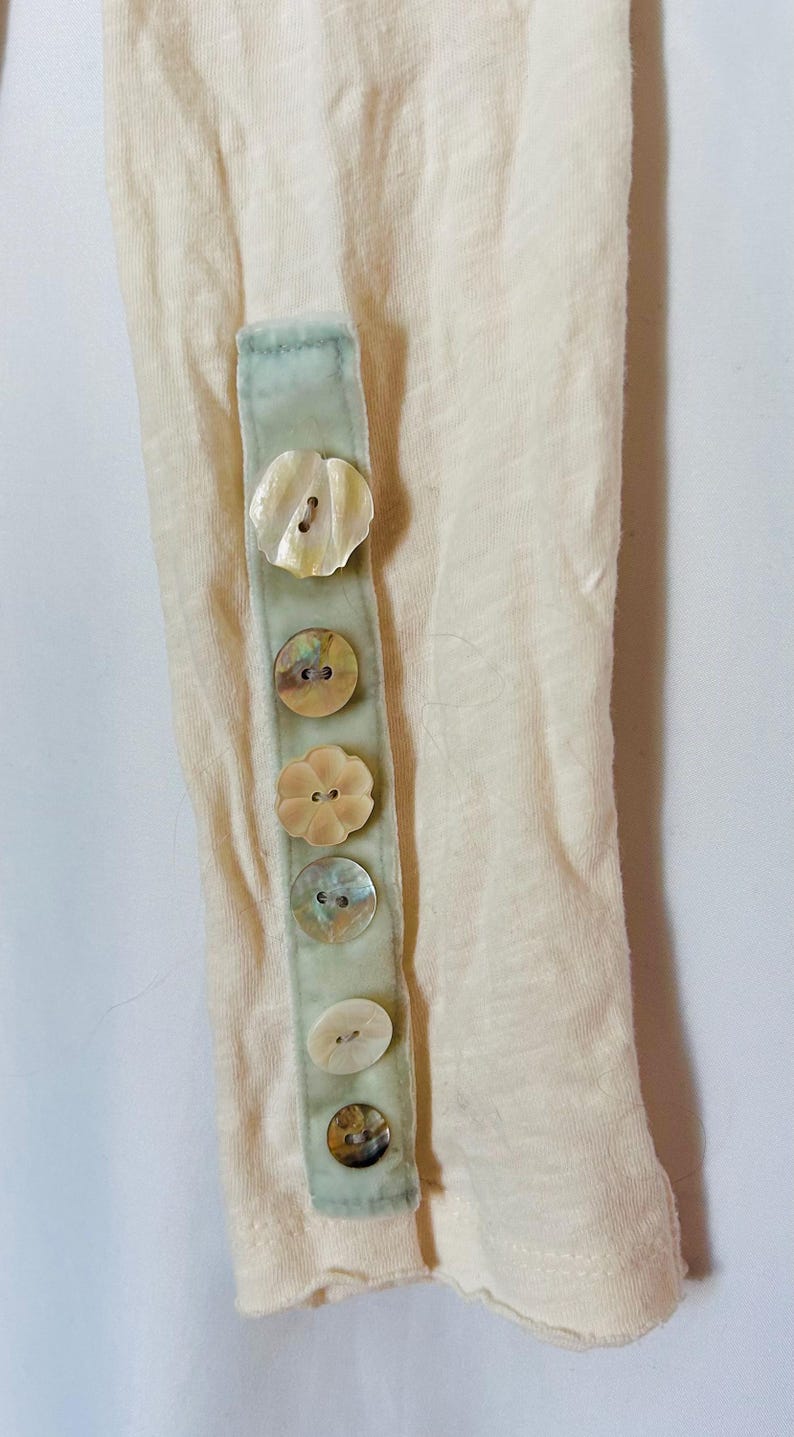 May include: Close-up of a cream-colored sleeve with a light green fabric strip. The strip is adorned with five iridescent shell buttons in various shapes. The sleeve's fabric has a slightly textured appearance.