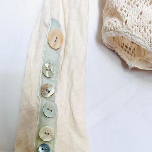 May include: Close-up of a cream-colored long-sleeved shirt with a row of iridescent buttons down the sleeve. The buttons are attached to a pale green ribbon. A portion of a cream-colored crocheted garment is visible.