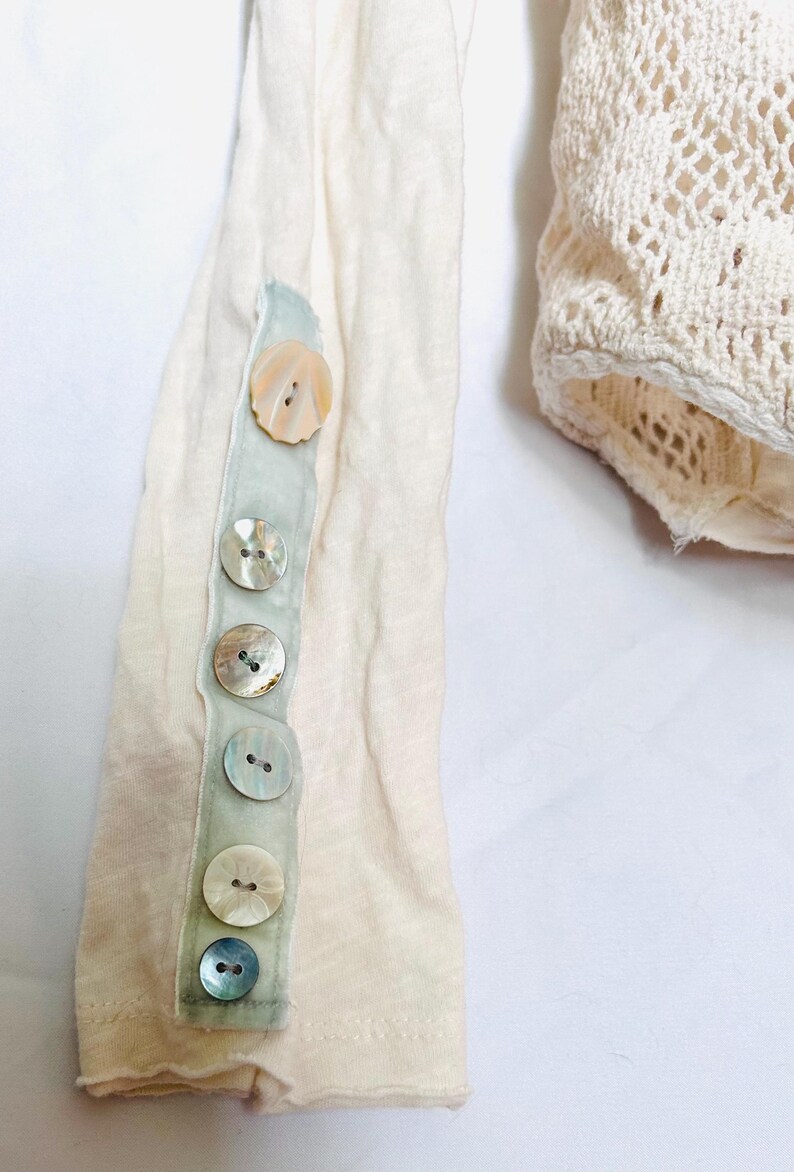 May include: Close-up of a cream-colored long-sleeved shirt with a row of iridescent buttons down the sleeve. The buttons are attached to a pale green ribbon. A portion of a cream-colored crocheted garment is visible.