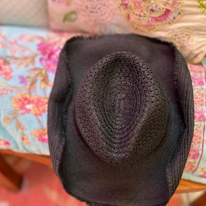 May include: A black woven straw hat with a wide brim. The hat has a textured crown and a slightly distressed edge. The hat is resting on a floral patterned surface.