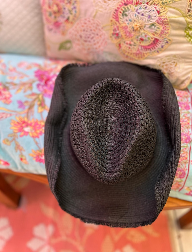 May include: A black woven straw hat with a wide brim. The hat has a textured crown and a slightly distressed edge. The hat is resting on a floral patterned surface.