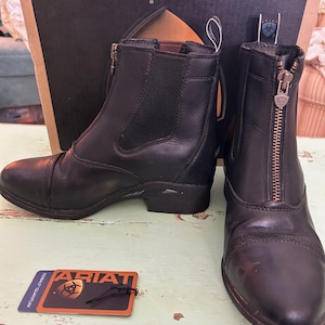 May include: A pair of black leather Ariat paddock boots featuring a front zipper closure with a branded pull tab, elastic side panels, and a rounded toe. A rectangular Ariat Cobalt Quantum brand tag is included with the footwear.