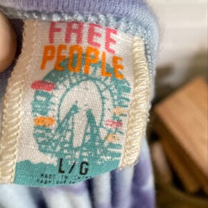 以下が含まれることがあります： ピンクとオレンジの文字で「Free People」と書かれた白いタグ。タグには、青と白の観覧車のグラフィックも描かれています。タグは、紫と青のタイダイ染めのシャツに付けられており、タグには「L/G」のサイズが印刷されています。