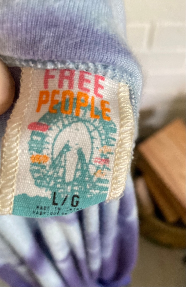 以下が含まれることがあります： ピンクとオレンジの文字で「Free People」と書かれた白いタグ。タグには、青と白の観覧車のグラフィックも描かれています。タグは、紫と青のタイダイ染めのシャツに付けられており、タグには「L/G」のサイズが印刷されています。