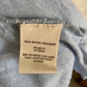 以下が含まれることがあります： 「100% RAYON/RAYONNE」、「RN 66170」、「CA 32054」、「SEE REVERSE FOR CARE」、「VOIR L'INVERSE POUR」、「LE SOIN」というテキストが書かれた薄いブルーのレーヨン生地のタグ。