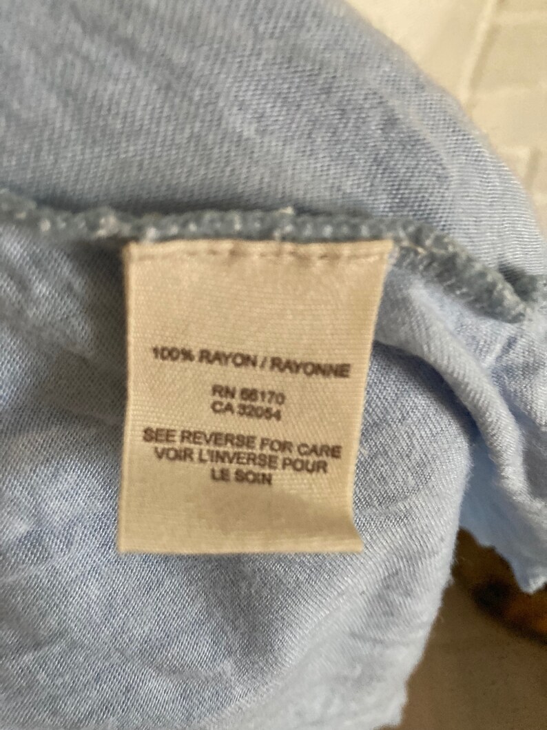 以下が含まれることがあります： 「100% RAYON/RAYONNE」、「RN 66170」、「CA 32054」、「SEE REVERSE FOR CARE」、「VOIR L'INVERSE POUR」、「LE SOIN」というテキストが書かれた薄いブルーのレーヨン生地のタグ。