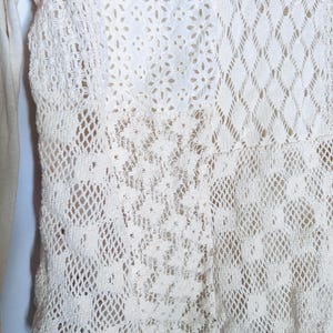 May include: Close-up of a cream-colored, open-weave garment with various lace patterns. The design includes floral, diamond, and geometric shapes, creating a textured and detailed appearance. The garment appears to be a top or a part of a dress.