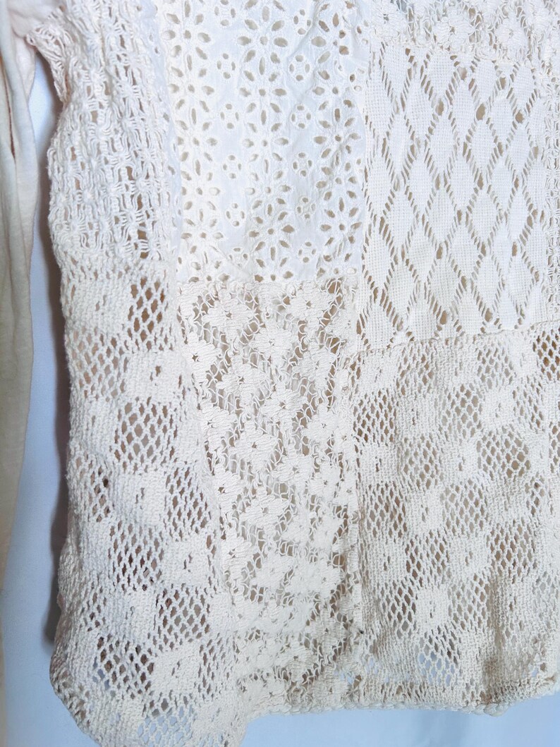 May include: Close-up of a cream-colored, open-weave garment with various lace patterns. The design includes floral, diamond, and geometric shapes, creating a textured and detailed appearance. The garment appears to be a top or a part of a dress.