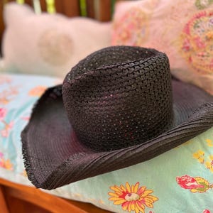 May include: A dark woven sun hat with a wide brim. The hat is made of a dark material and has a structured crown. The hat is resting on a floral patterned surface, suggesting outdoor or summer wear.