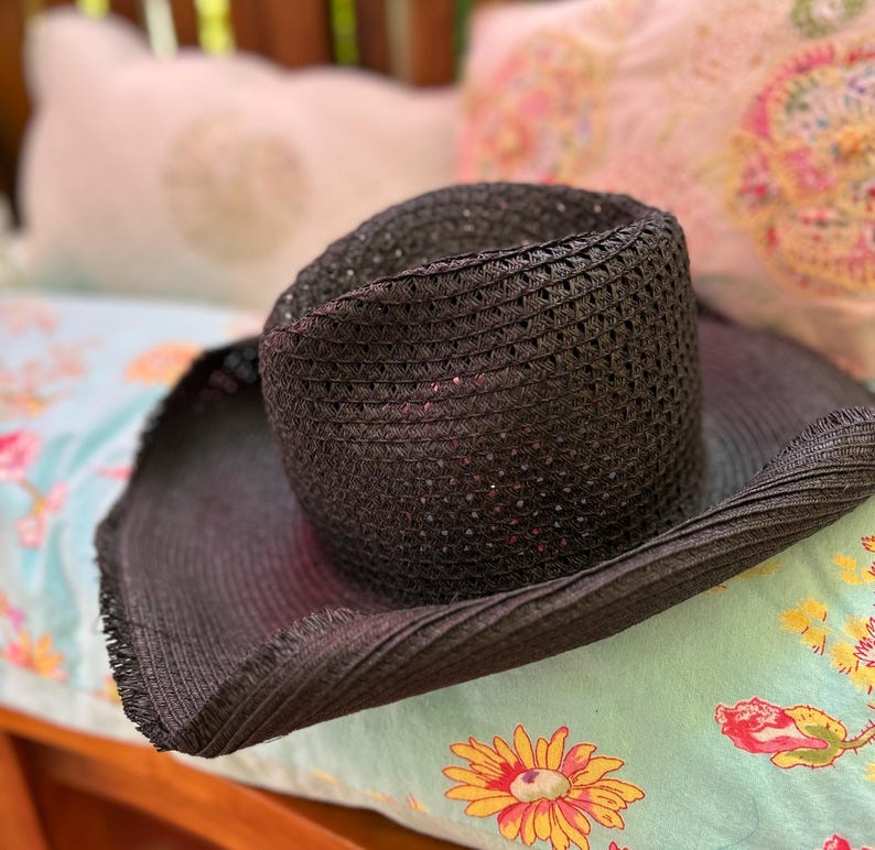 May include: A dark woven sun hat with a wide brim. The hat is made of a dark material and has a structured crown. The hat is resting on a floral patterned surface, suggesting outdoor or summer wear.