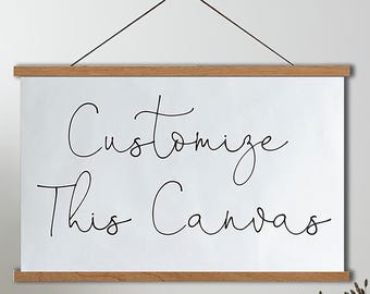 Op de afbeelding: Een wit rechthoekig canvas hangend aan twee horizontale houten latten met een dun zwart touwtje. Het canvas bevat de zwarte cursieve tekst "Customize This Canvas" gecentreerd op het oppervlak. Het ontwerp is aan één punt opgehangen.